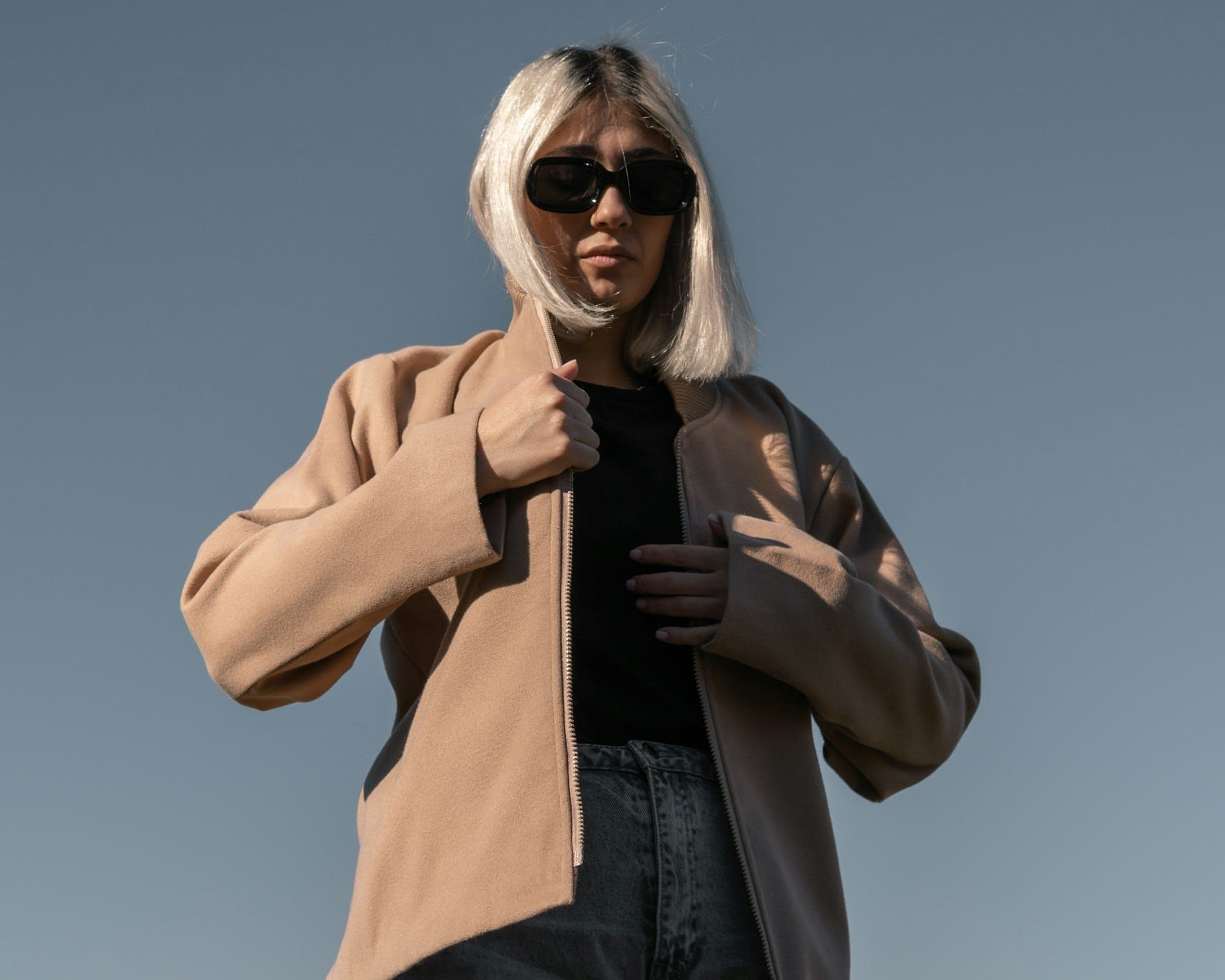 Model in black oval sunglasses and a camel bomber jacket, shot from below against the sky